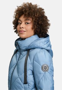 Jasnoniebieska pikowana kurtka z wysokim kołnierzem i kapturem, z czarnymi błyszczącymi paskami i naszywką z logo na lewym ramieniu. Miękka w dotyku.