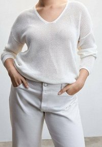 Weißer, locker gestrickter Pullover mit tiefem V-Ausschnitt und langen Ärmeln, kombiniert mit hochgeschnittenen, hellen, weit geschnittenen Hosen mit Frontknopf.