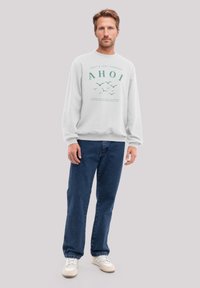 Weißes Sweatshirt mit grünem Druck und dem Text "AHOI" sowie Vögeln. Kombiniert mit blauen Jeans und weißen Sneakers. Lässiger Schnitt.