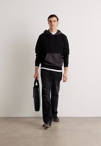 Felpa nera con tasca grigia, t-shirt bianca sotto, pantaloni neri e sneaker grigi. Tiene una borsa nera mentre cammina su uno sfondo neutro.