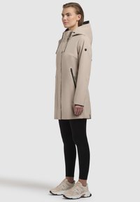 Manteau imperméable beige avec capuche, fermeture éclair avant, accents noirs et deux poches latérales zippées, associé à un legging noir et des baskets beiges.
