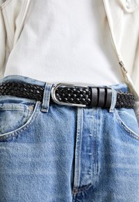 Fekete, fonott bőr öv ezüst színű fém csattal, világoskék farmernadrágra és fehér, bordázott felsőre húzva.