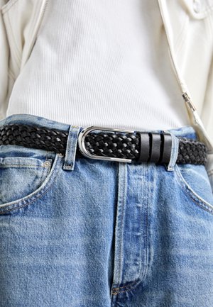 Curea din piele împletită neagră cu o cataramă metalică argintie, purtată peste blugi de denim albastru deschis și un tricou alb cu nervuri.
