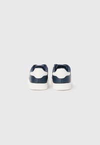 Ein Paar marineblaue und weiße Low-Top-Sneaker von hinten vor einem schlichten hellgrauen Hintergrund gezeigt.