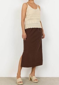 Vrouw in een beige gehaakte mouwloze top, een bruine midi-rok met een zijsplit en beige plateau-sandalen, staande op een betonvloer.