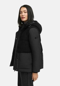 Black puffer jacket with a textured fleece upper section, hood, and snap-button front. It features a side pocket and is paired with black jeans.