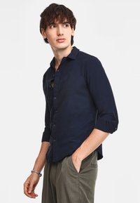 Camicia blu navy con bottoni realizzata in un tessuto morbido, con collo a camicia, maniche lunghe con risvolti e orlo dritto. Abbinata a pantaloni oliva.