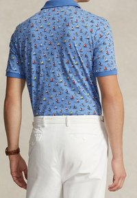 Ljusblå polotröja med ett mönster av färgglada segelbåtar. Korta ärmar, ribbad krage och en slät textur. Kombineras med vita byxor.