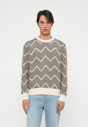 Gebreide trui in grijs en crème met een chevronpatroon, ronde hals, ribgebreide manchetten en zoom, gedragen over lichtblauwe jeans.