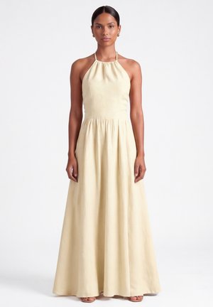 Kobieta stoi w bezowej, bezrękawnej sukience na ramiączkach sięgającej do ziemi z marszczonym gorsetem na jednolitym białym tle.