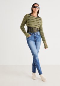 Suéter corto de punto en verde oliva con tejido abierto, combinado con jeans de tiro alto azules y botas de tobillo beige, llevado con gafas de sol oscuras.