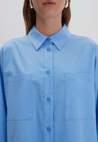 Camicia azzurra chiara con bottone in tessuto liscio, dotata di colletto, due tasche frontali e sei bottoni abbinati lungo la parte anteriore.