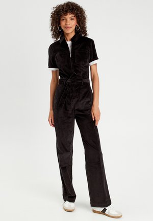 Mono de pana negra con mangas cortas, cierre de cremallera, dos bolsillos en el pecho y un lazo en la cintura. Combinado con zapatillas blancas y pantalones de corte relajado.
