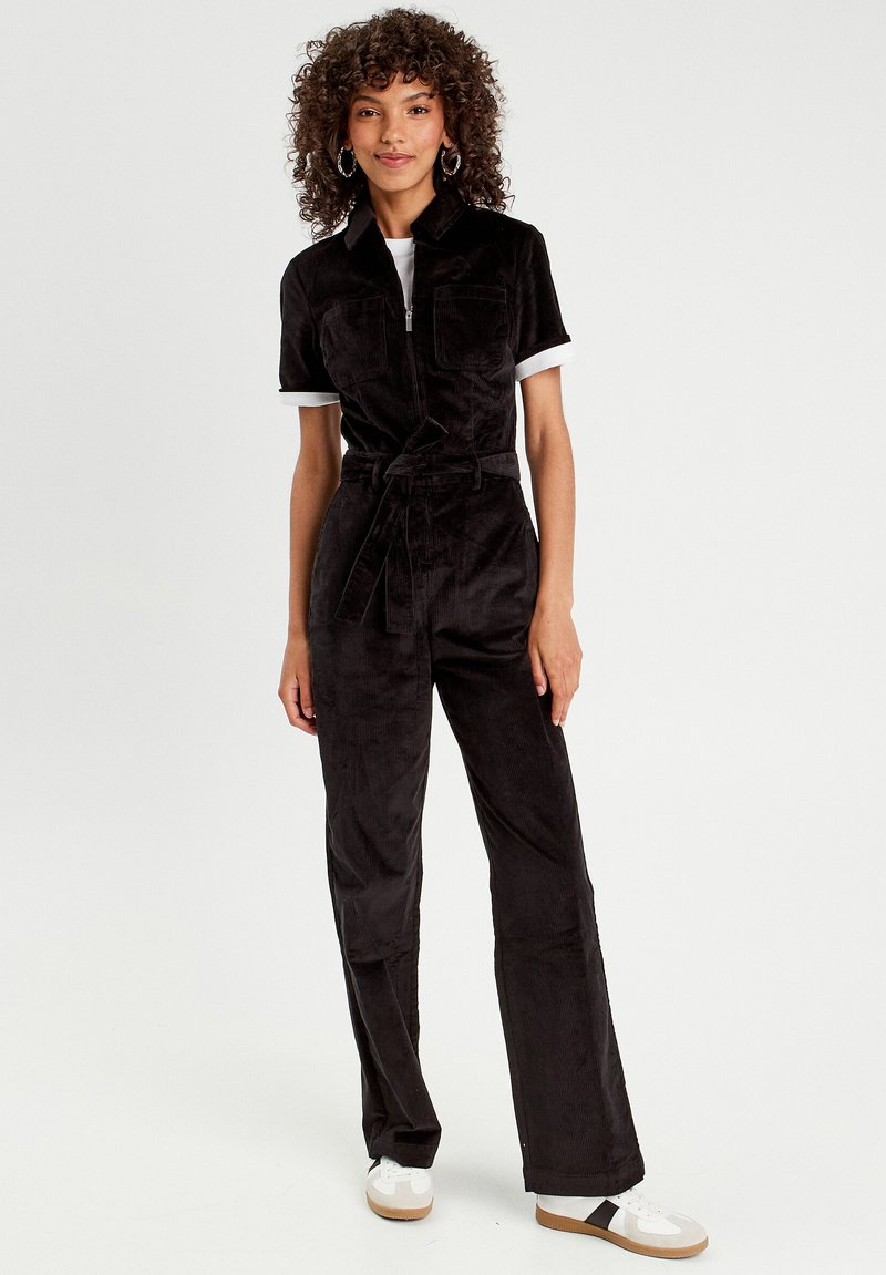 Black corduroy jumpsuit with short sleeves, zip closure, two chest pockets, and a waist tie. Paired with white sneakers and relaxed-fit pants.