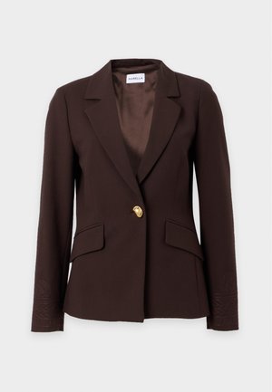 Une femme porte un blazer marron foncé, un haut bleu marine, un pantalon beige à jambes larges et des chaussures marron sur un fond clair uni.