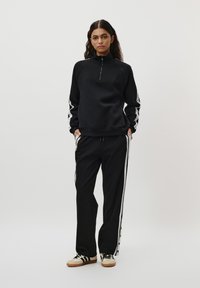 Svart fleece-genser med halv glidelås og raglanermer. Svart bukse med hvite sidestreker. Begge har en avslappet passform.