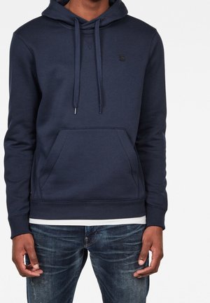 Mand iført en mørkeblå hættetrøje med snører og frontlomme, kombineret med blå denimjeans og en hvid skjorte under.