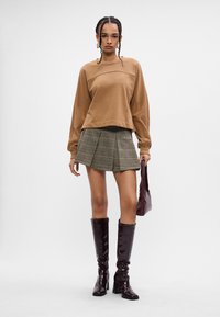 Sudadera scurtă maro, fustă mini în carouri, cizme lucioase maro până la genunchi și o geantă asortată. Materiale texturate și design ajustat.