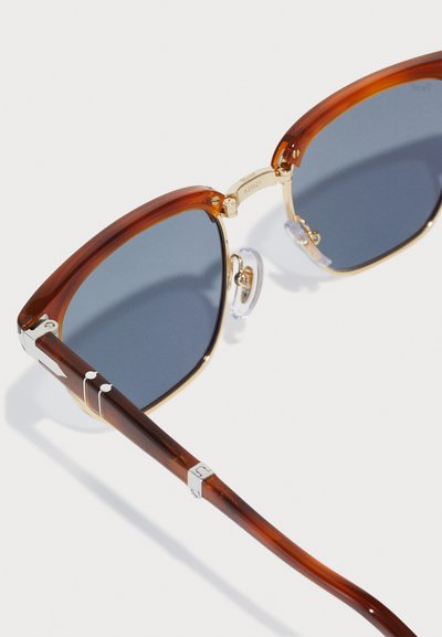 Ochelari de soare cu nuanțe de brown și accente aurii, cu lentile rectangulare și un suport de nas transparent. Brațul are o finisare lucioasă în nuanță de brown, cu detalii gravate.