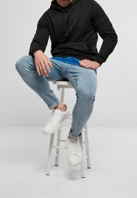 Czarna bluza z kapturem, niebieska koszulka, jasnoniebieskie wąskie dżinsy i białe sneakersy. Siedząca na białym stołku na jasnym tle.