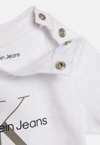 Body de bebé de algodón blanco con broches de presión plateados en el hombro y un logotipo impreso, que presenta una gran "K" estilizada y el nombre de la marca.