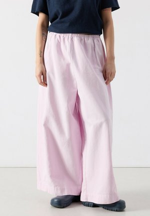 Person trägt weit geschnittene, hellpinkfarbene Hosen mit weitem Bein, schwarze Schuhe und ein dunkelblaues Oberteil und steht vor einem einfarbigen Hintergrund.