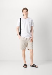 Tricou alb cu logo brodat, pantaloni scurți bej și sandale negre tip slide. Design simplu, croială casual, material moale și accente minime.