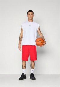 Valge varrukateta särk musta Nike logoga, punased lühikesed püksid, mustad kingad, hoiab korvpalli. Käte peal tätoveeringud ja mustrilised sokid.