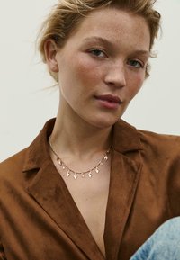 Collana in oro rosa con pendenti in cristallo trasparente, indossata sopra una giacca in suede marrone. Lo sfondo è neutro e minimal.