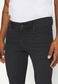 Tmavě modré džíny z denimu s úzkým střihem. Mají zapínání na knoflíky, přední kapsy a jemné švy. Materiál má hladkou texturu.