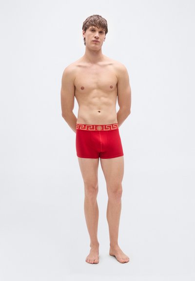 Κόκκινα ανδρικά σορτς από μαλακό ύφασμα με χρυσή ζώνη μέσης διακοσμημένη με μοτίβο ελληνικού κλειδιού. Τα σορτς είναι εφαρμοστά και έχουν μήκος μέχρι τη μέση του μηρού.