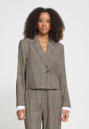 Jeune femme aux cheveux bouclés portant un blazer court à carreaux gris et un pantalon taille haute assorti, debout devant un fond uni.