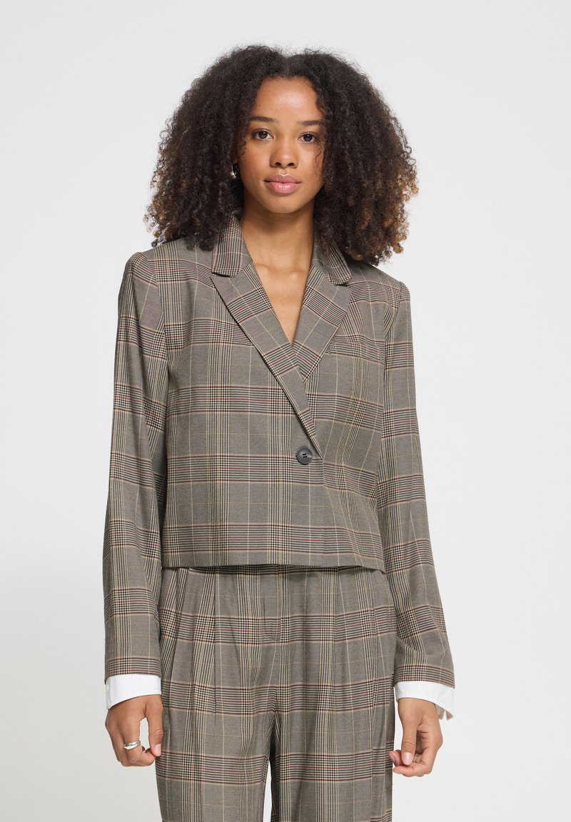 Jeune femme aux cheveux bouclés portant un blazer court à carreaux gris et un pantalon taille haute assorti, debout devant un fond uni.