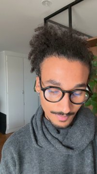 Jeune homme aux cheveux bouclés, portant des lunettes et des boucles d'oreilles, regarde vers le bas, vêtu d'un pull gris foncé dans une pièce lumineuse avec une plante et une armoire.
