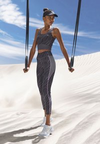 Schwarz-graues Sport-BH und Leggings mit pinken Akzenten, kombiniert mit weißen Sportschuhen, vor einem sandigen Hintergrund.