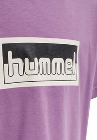Fioletowa bawełniana koszulka z krótkim rękawem, z okrągłym dekoltem, z białą prostokątną grafiką i czarnym napisem „hummel” w pogrubionej, zaokrąglonej czcionce.