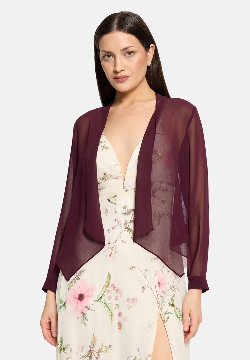Femme aux longs cheveux foncés portant une veste violette transparente ouverte sur une robe blanche fleurie avec un profond décolleté en V et une fente à la cuisse.