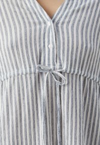 Svītraina blouse zilos un baltos toņos, ar V veida kakla izgriezumu, pogu aizdari un auklu pievilkšanu jostasvietā, izgatavota no teksturēta auduma.