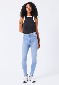 Zwarte mouwloze top met witte accenten, hoog-waist lichtblauwe skinny jeans en witte sneakers met donkere accenten. Krullend haar.