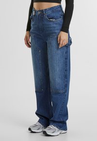 Locker sitzende blaue Jeans aus Denim mit hoher Taille, großen Fronttaschen und kontrastierenden Paneelen, kombiniert mit grauen Sportschuhen.