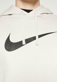 Person i en vit huvtröja med en stor svart Nike swoosh-logotyp och vita dragband, sedd från halsen till bröstet.