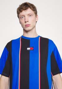 Tricou cu dungi albastre, negre și roz, fabricat din bumbac. Prezintă o pată rectangulată cu logo-ul Tommy Jeans în centru. Mâneci scurte.