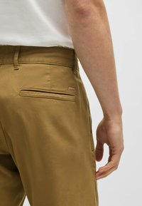 Khaki trousers made of cotton, featuring a smooth texture, side and back pockets, belt loops, and a small brand label on the back pocket.