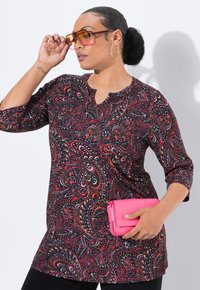 Vrouw in een gestreepte tuniek, grote oranjeachtige zonnebril, gouden creolen, houdend een felroze clutch tegen een witte achtergrond.