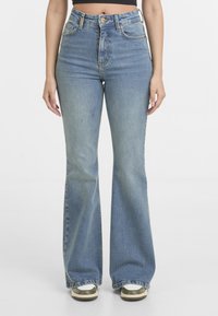 Femme portant un jean flare bleu taille haute et des baskets blanches, debout devant un fond blanc uni.