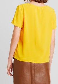 Blouse jaune à manches courtes en tissu lisse, dotée d'un col arrondi et de petites fentes latérales, associée à une jupe marron texturée.