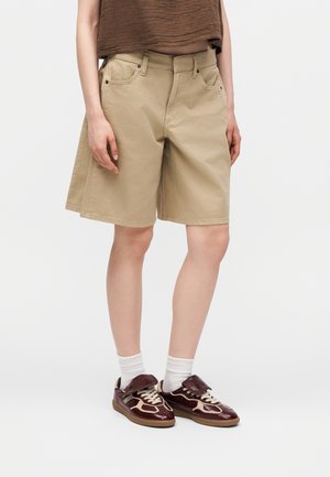 Osoba nosząca beżowe szorty do kolan, brązowy krótki top, białe skarpetki i bordowo-beżowe sneakersy stojąca na jednolitym tle.