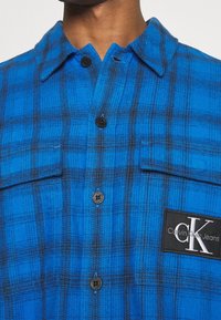 Blue plaid shirt made of flannel fabric featuring black accents, two chest pockets, and a logo patch from Calvin Klein Jeans.