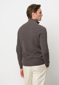 Enganliegender grauer Rippstrickpullover mit hohem Kragen und langen Ärmeln, versehen mit einem strukturierten Strickmuster und einem geraden Saum, getragen über einer hellen Hose.