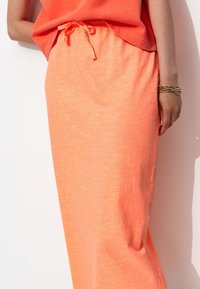 Jupe longue corail-orange en tissu léger avec une taille à cordon. Présente une coupe décontractée et une texture subtile.